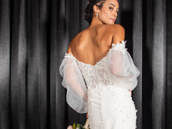 Off the shoulder lace bodice wedding dress with sheer sleeves modeled by a bride in front of black curtains.