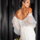 Off the shoulder lace bodice wedding dress with sheer sleeves modeled by a bride in front of black curtains.