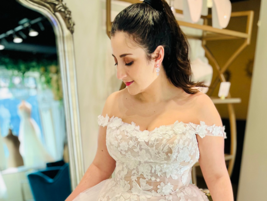 Beautiful off-the-shoulder blush lace floral wedding gown, worn by a brunette model at The Wedding Shoppe in Berkley, Michigan.