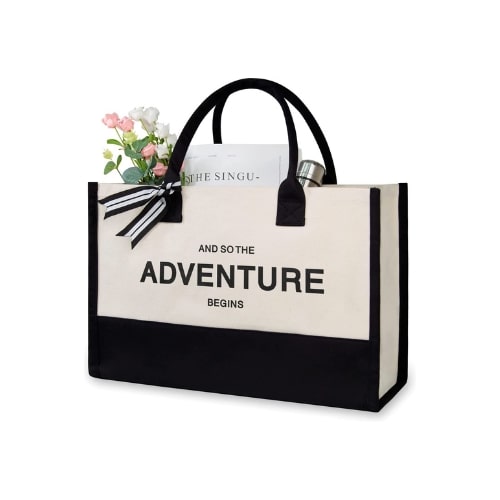 Black and beige, rectangular canvas bag. The bottom half of the bag is black while the beige top half features "And So The Adventure Begins" written in bold, black font.