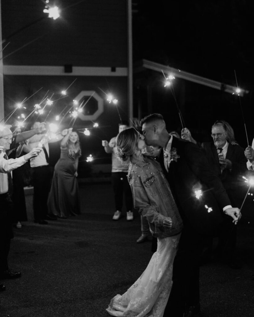 black and white photography of married couple kissing with partygoers holding sparklers