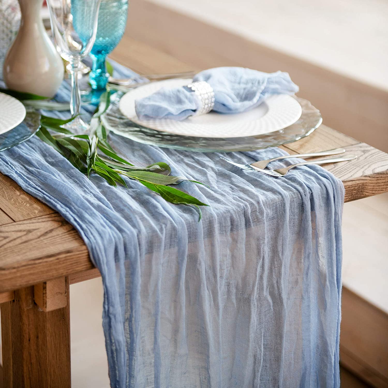 Wedding Reception table runner made of sea blue cheesecloth. On the runner is a table dinner setting and an assortment of glasses can be seen nearby.