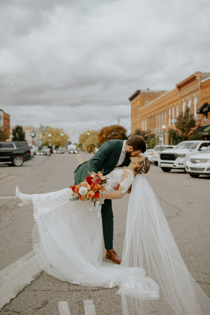 married couple kissing on the street with the bride holding an assortment of red, orange, and white flowers