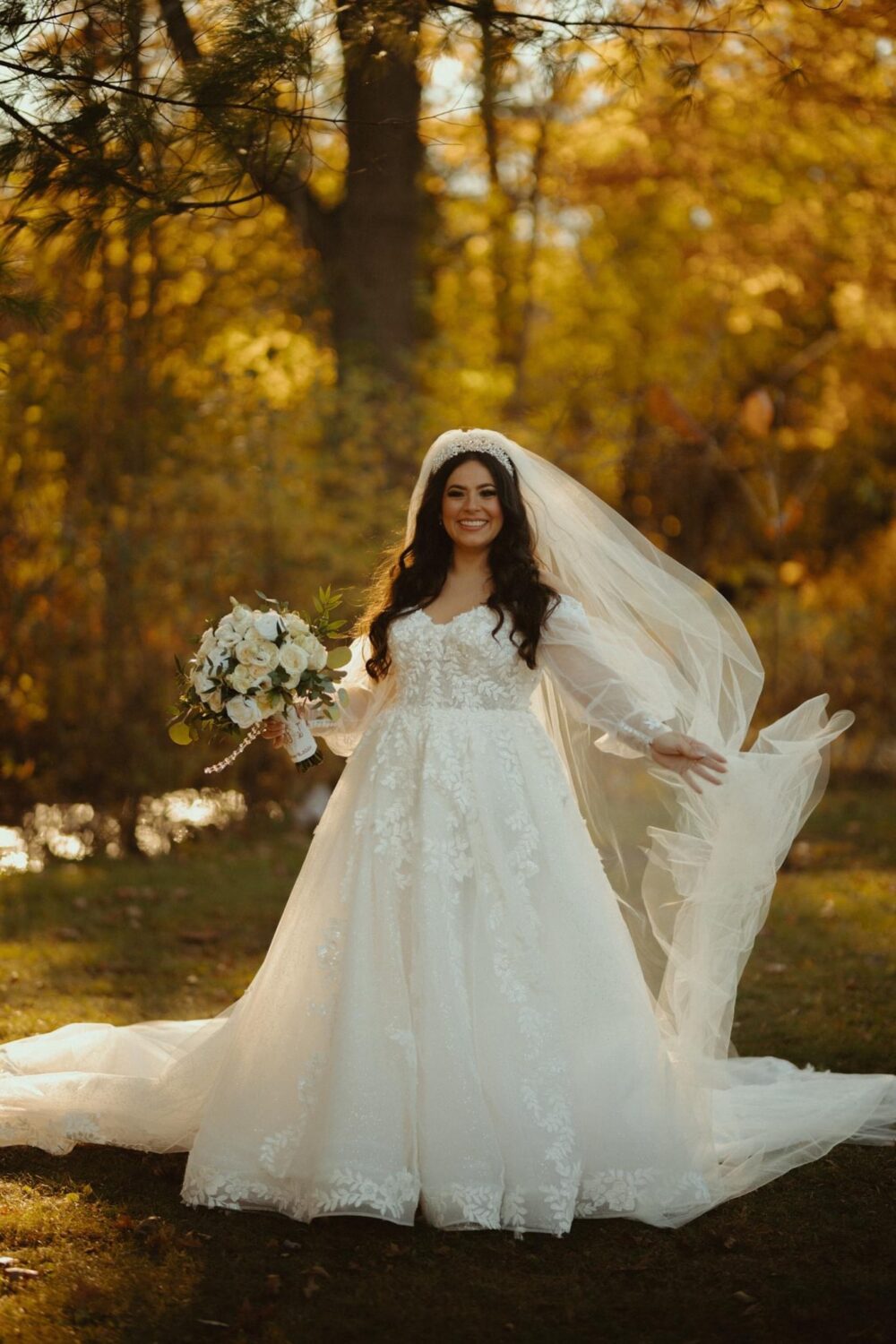Bride stands smiling in an outdoor fall setting holding a bouquet of white flowers.