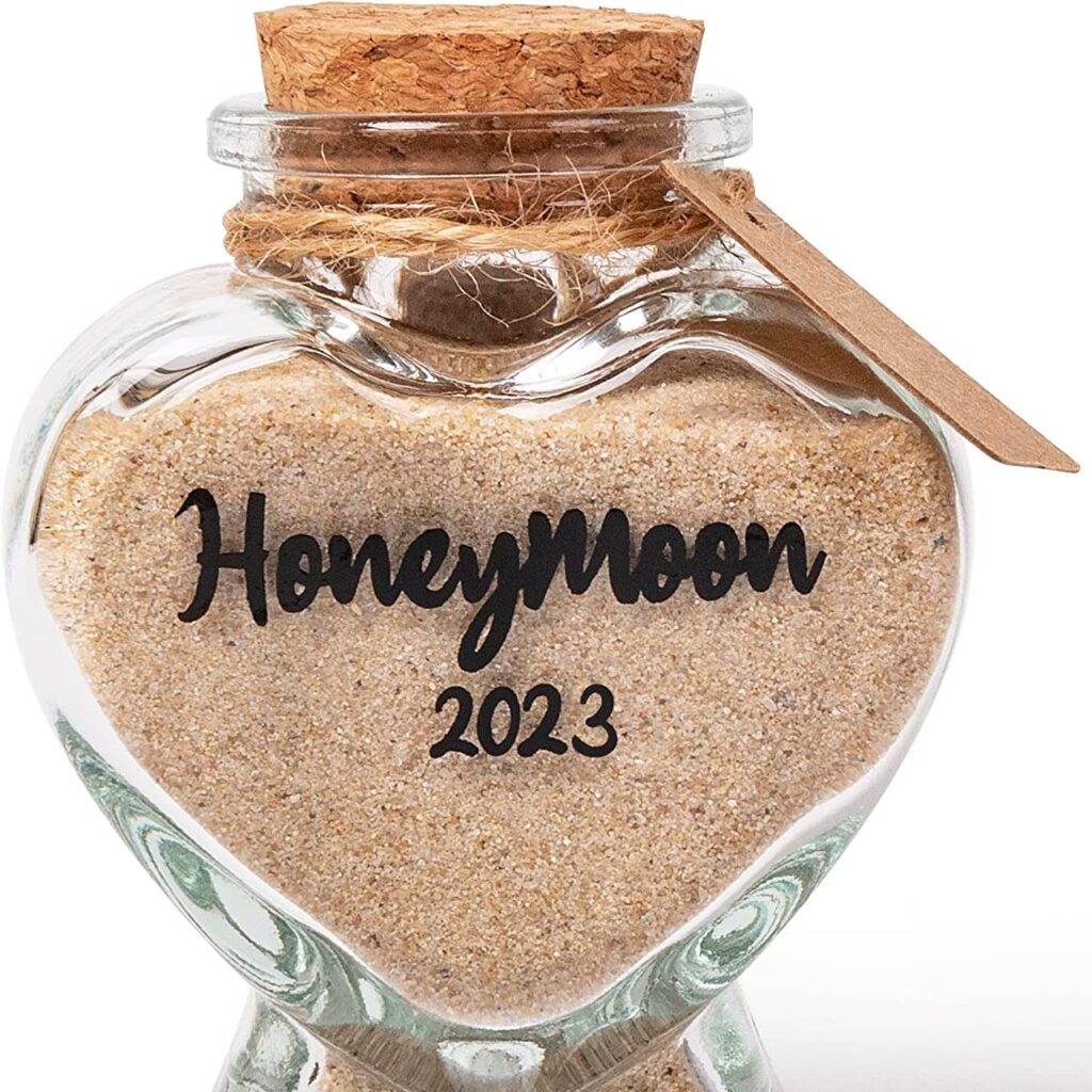 Small, heart shaped glass with cork stopper and twine around the neck. The glass is filled with sand and says "honeymoon 2023"