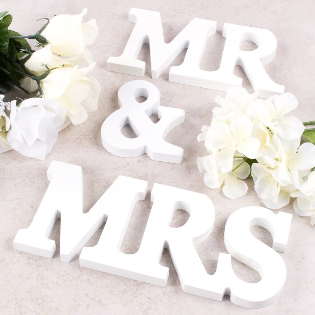 3D, white wooden pieces spelling out "Mr & Mrs" laid out on the floor. Nearby are white flowers.