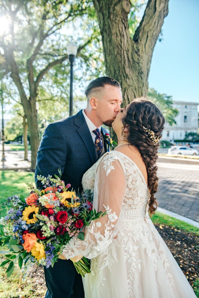 married couple kissing outdoors while the bride holds an assortment of sunflowers and other flowers