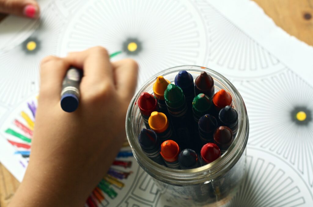 child coloring on a coloring page with a blue crayon with a jar of other crayons nearby