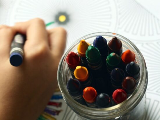 child coloring on a coloring page with a blue crayon with a jar of other crayons nearby