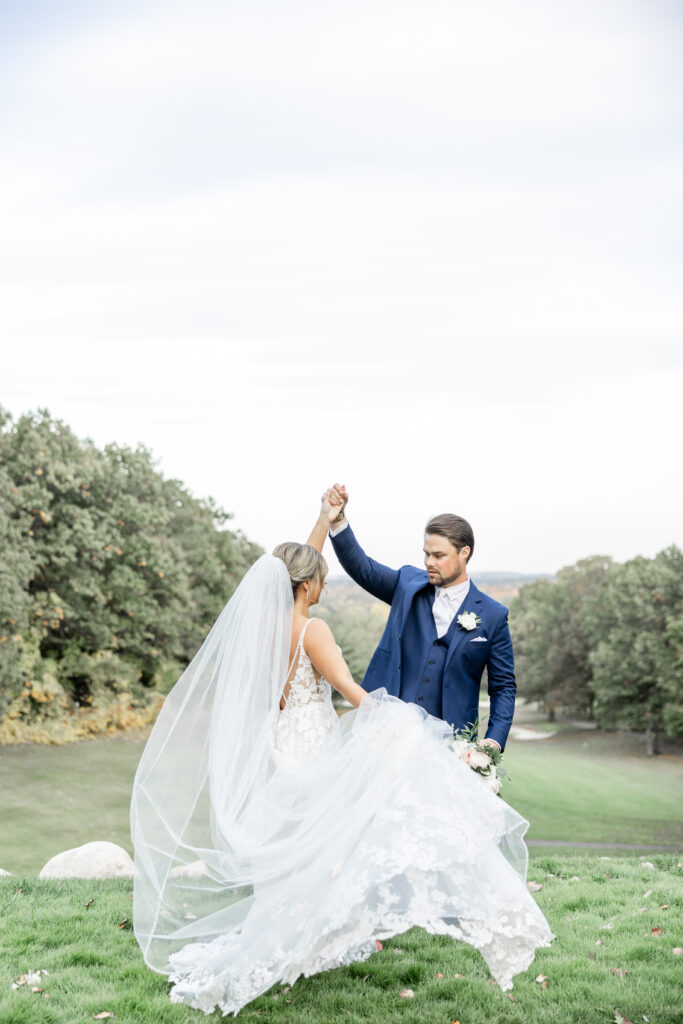 Groom in blue suit spinning his bride in front of him. His bride is twirling and holding her long wedding dress and viel.