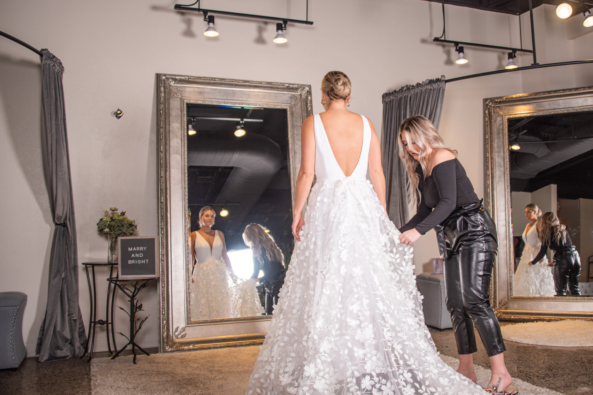 Bride-to-be standing in front of The Wedding Shoppe's mirror admiring her wedding dress while her bridal stylist adjusts the train.