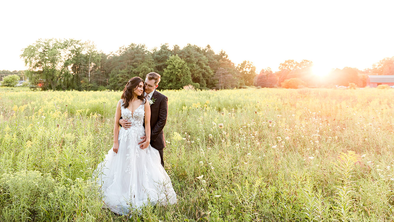 Bride and Groom standing together in a field of wildflowers. The groom is embracing his bride from behind while she looks off into the sunset.