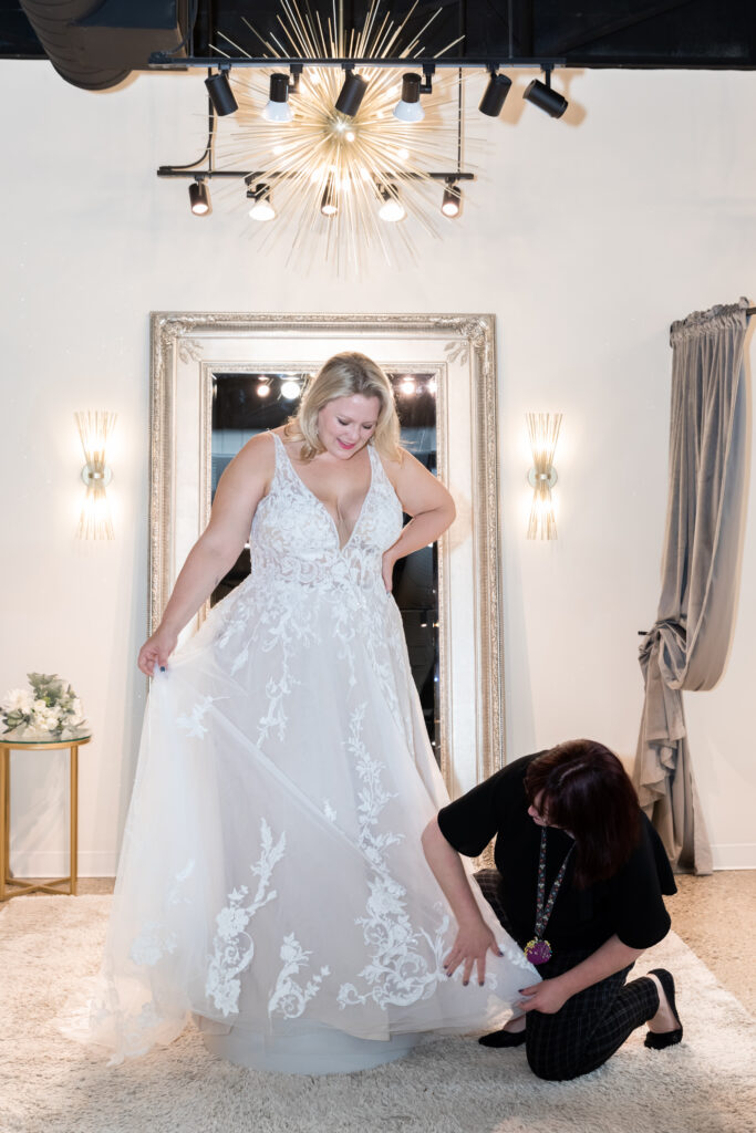 Bride-to-Be standing on a pedestal in The Wedding Shoppe while her alterations seamstress adjusts her skirt.