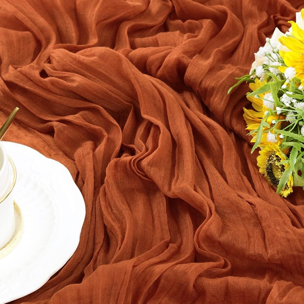 Cheesecloth table runner in terracotta color. On the side is a small bouquet of yellow and white flowers.