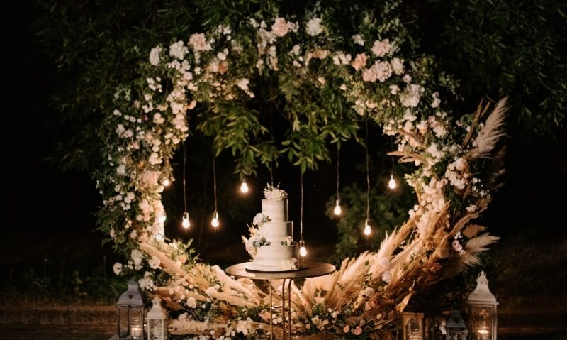 floral and plant archway with hanging lights behind a tiered wedding cake
