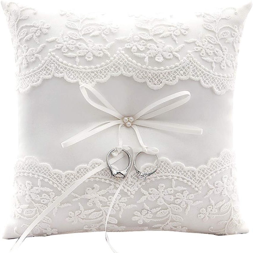 Small and white, square pillow with lace embroidery on the top and bottom. in the middle is a bow to tie the wedding rings to.