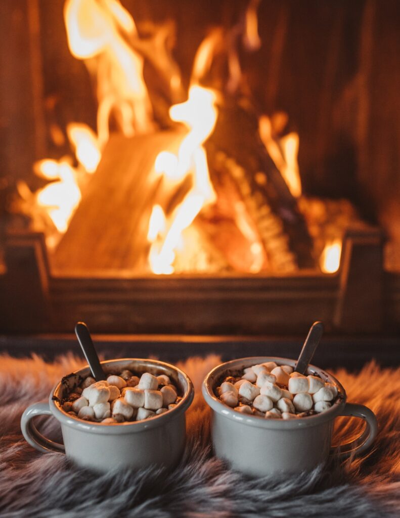 two mugs of hot chocolate and marshmallows in front of a roaring log fire