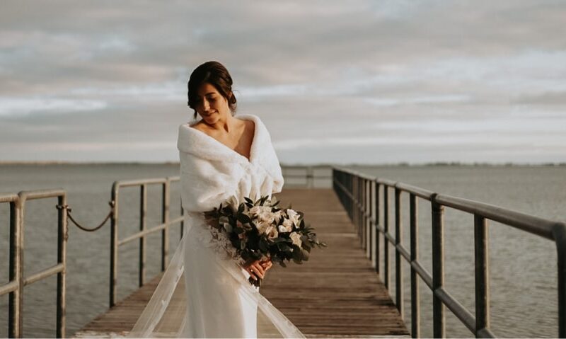 a bride in a dress with a wrap holding a bouquet of flowers on a pier