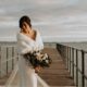 a bride in a dress with a wrap holding a bouquet of flowers on a pier
