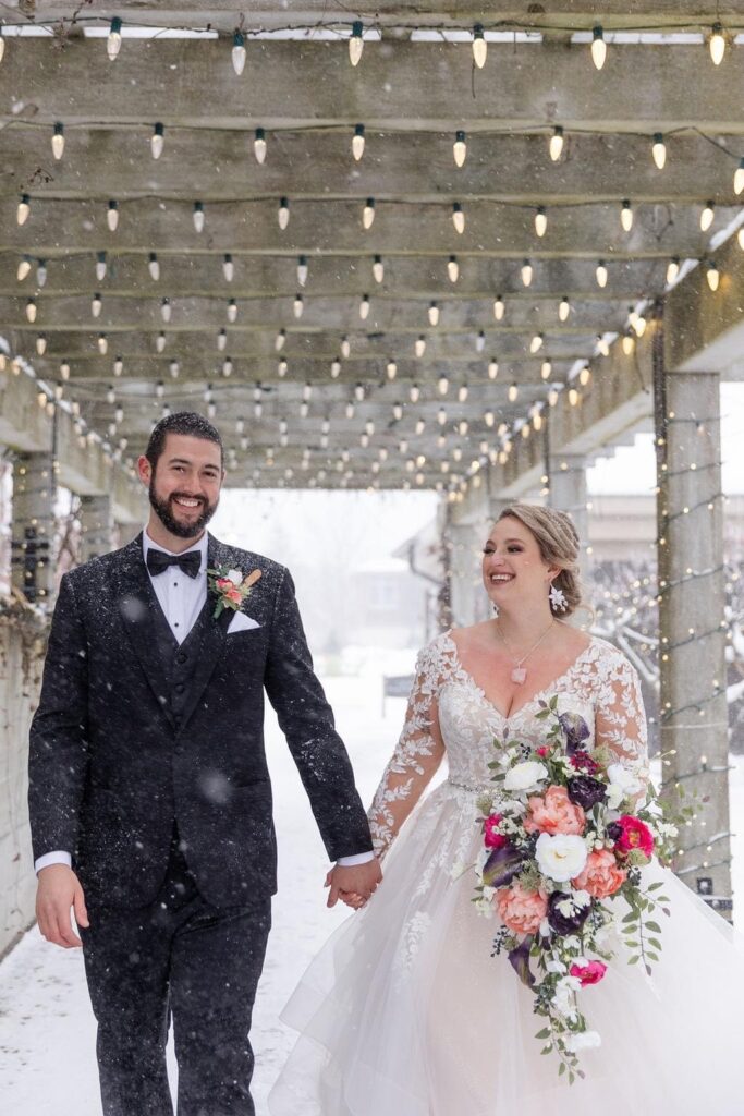 bride and groom walking outside as snow falls around them. The bride holds a colorful bouquet of flowers.