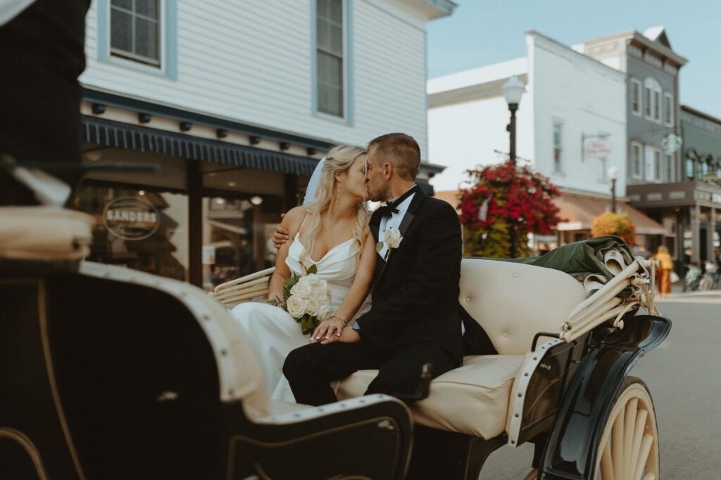 newly married couple kissing as they ride off on a horse-drawn carriage