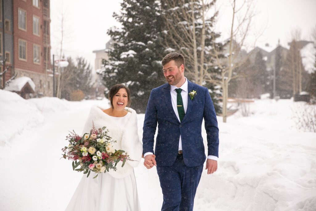 bride and groom walking in a winter wonderland holding a bouquet of colorful flowers