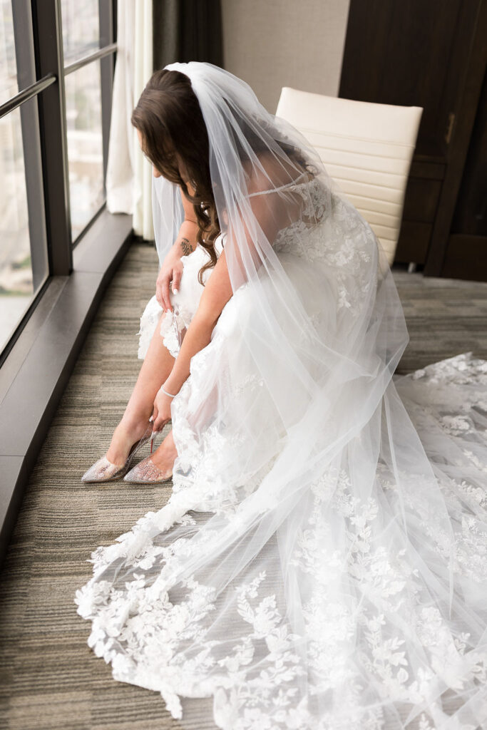 The Wedding Shoppe bride seated and putting on her shoes while wearing her wedding dress. Her dress and veil are spread out around her chair.