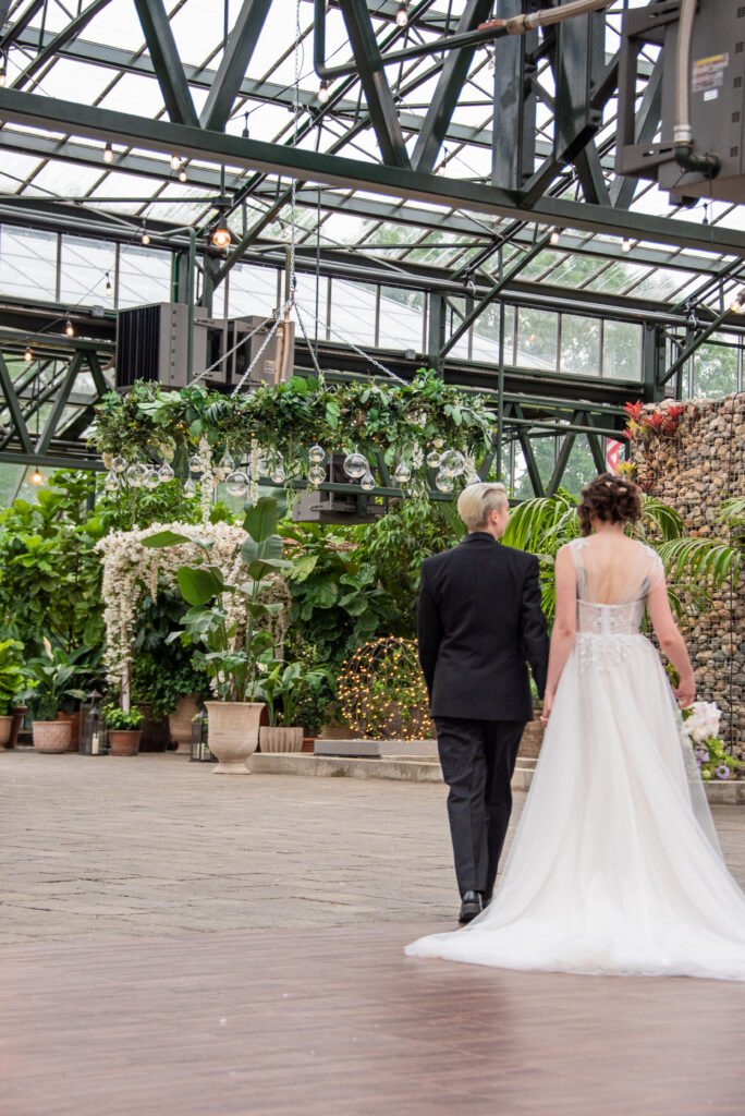 Corinne and Alicia at the Planterra Conservatory, wearing the Allure dress and the Randazzo Formalwear suit. They are holding hands and walking through the conservatory