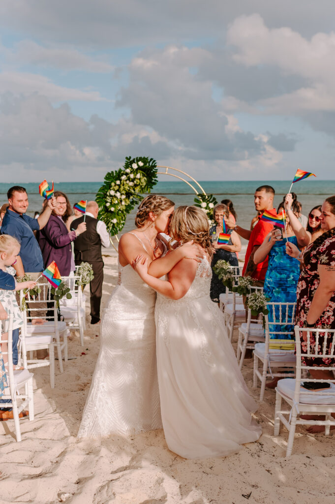 brides kissing at their sunset beach wedding surrounded by friends and family