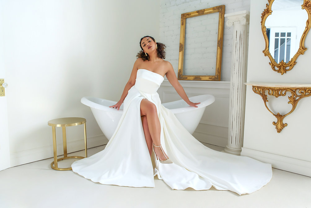 Stunning bride wearing her wedding dress and sitting on the edge of a clawfoot bathtub. In the bathroom around her are golden accent decor pieces. 