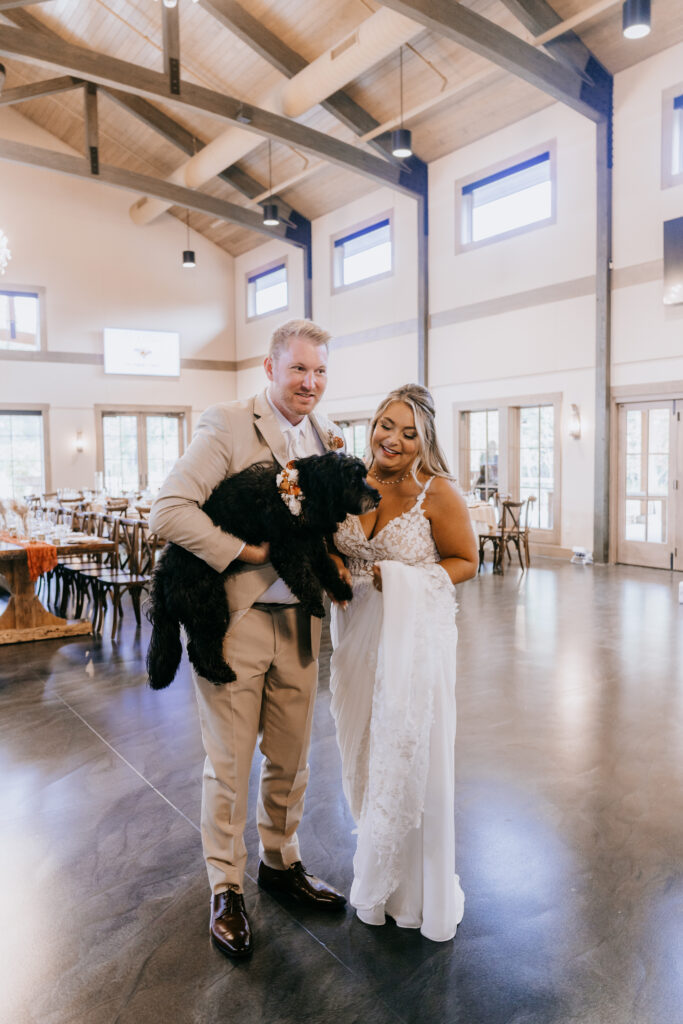 A newly married bride and groom stand holding their black dog at their wedding