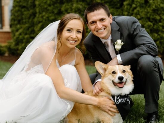 A husband and wife with their corgi dog who wears a "best boy" scarf