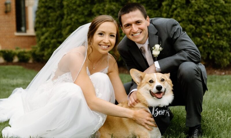 A husband and wife with their corgi dog who wears a "best boy" scarf