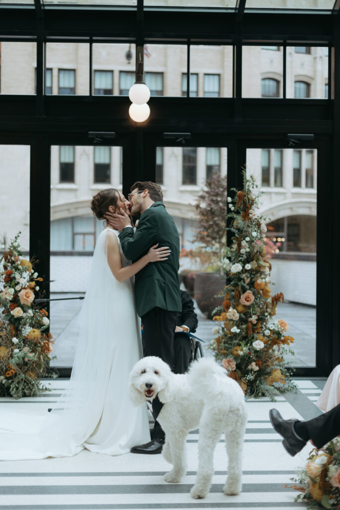 husband and wife sharing a kiss at their wedding as their white dog looks at the camera