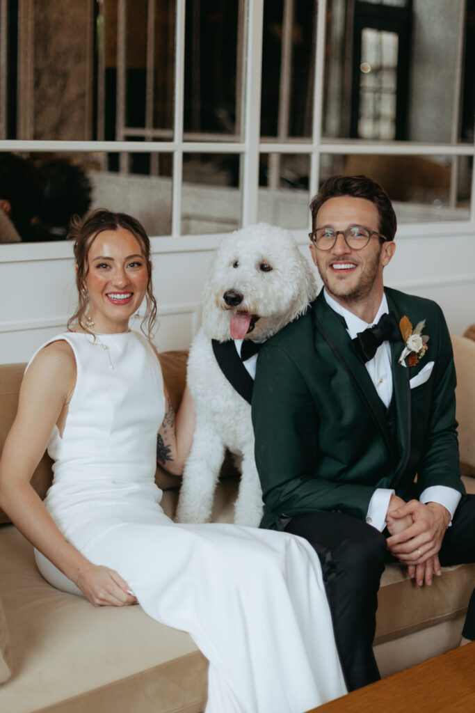 husband and wife sitting on a couch at their wedding with their white dog