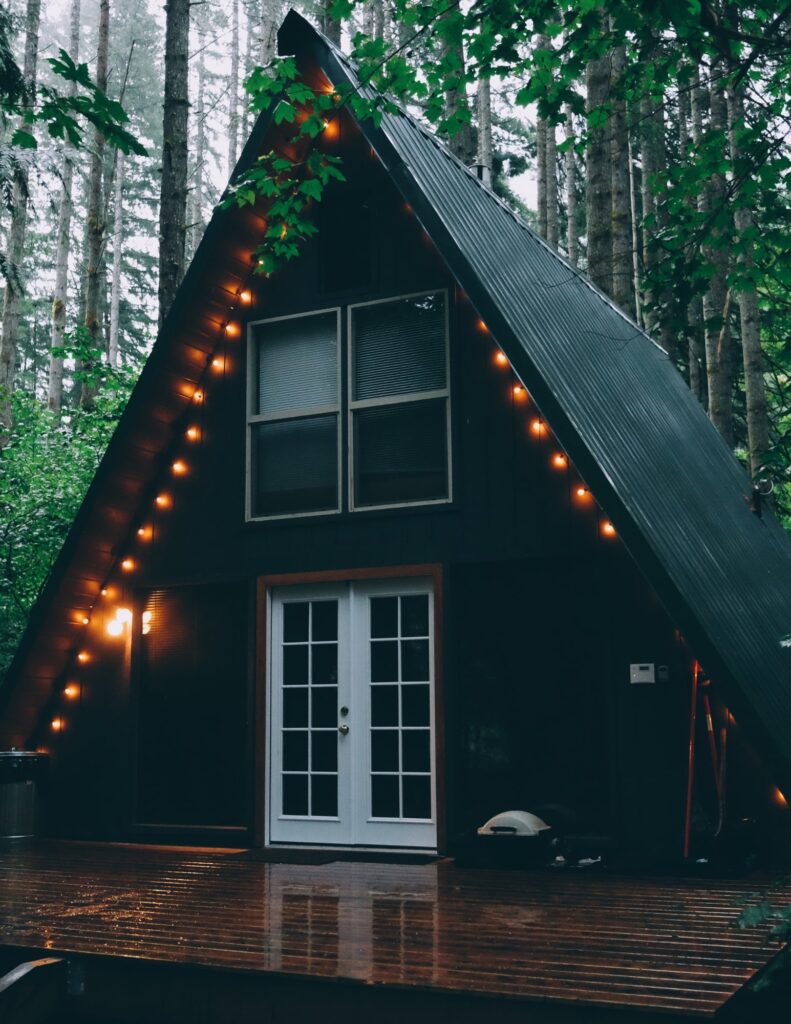 A simple A-shaped cabin painted black and adorned with string lights in the middle of the woods.