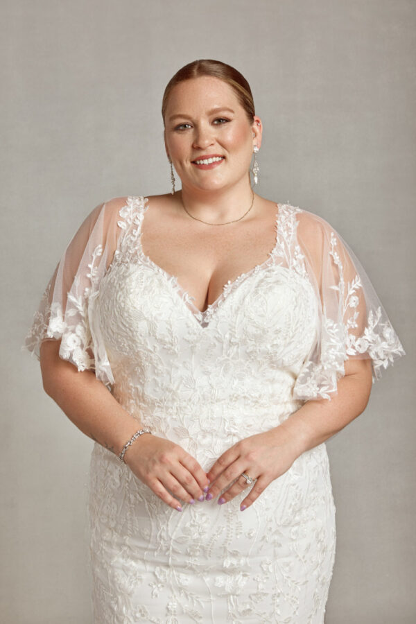 Curvy bride to be wearing a wedding dress with sleeves. The dress has small floral detailing on the bodice, sleeves and skirt.