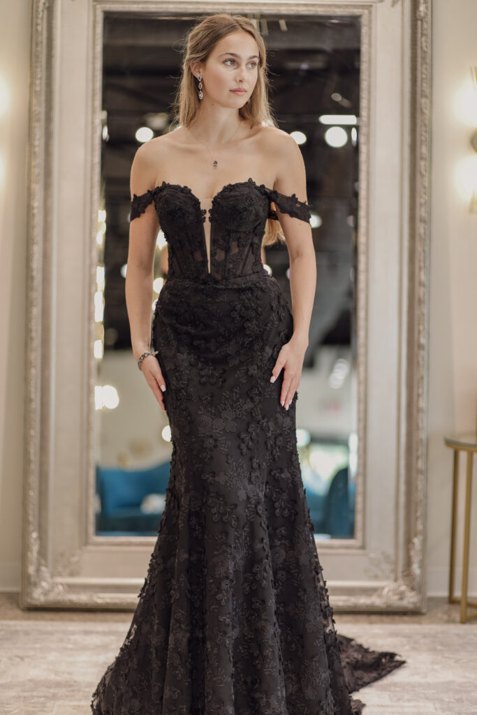 A bride wearing a black wedding dress with straps on the arms, in front of a mirror at a Michigan wedding shop.