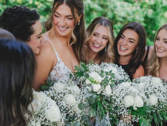 A group of smiling bridesmaids around the bride, each holding a bouquet of white flowers with green details.