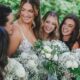 A group of smiling bridesmaids around the bride, each holding a bouquet of white flowers with green details.