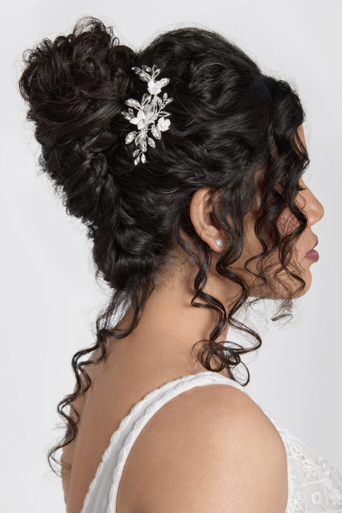 A side profile of a bride with curly dark hair styled in an elegant updo, adorned with a silver floral hairpin. The hairpin adds a sparkling accent to her sophisticated hairstyle.