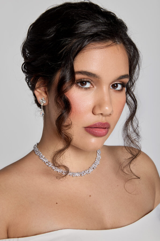 A close-up of a bride wearing a silver necklace and matching earrings. Her hair is styled in a classic updo, and she has a confident and poised expression.