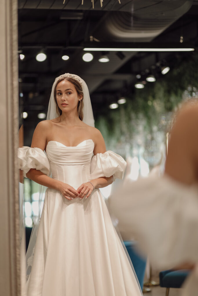 A bride wearing an off-the-shoulder wedding dress with voluminous sleeves and a long veil, standing in a softly lit room. The dress features a structured bodice with elegant folds.