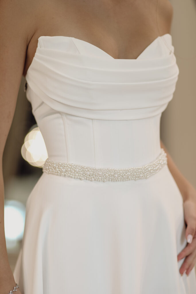 A bride wearing a white wedding dress with a pearl-beaded belt. The close-up focuses on the dress's structured bodice and the elegant pearl belt.