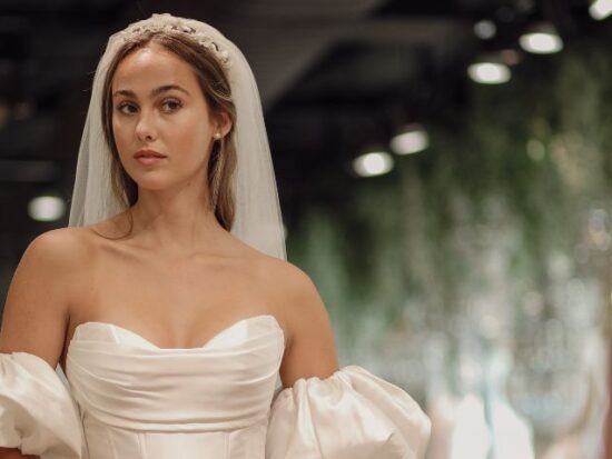 A bride wearing an off-the-shoulder wedding dress with voluminous sleeves and a long veil, standing in a softly lit room. The dress features a structured bodice with elegant folds.