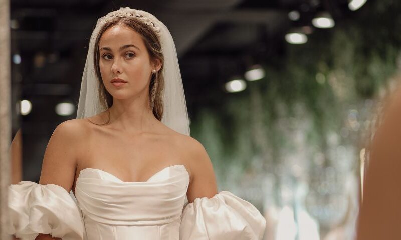 A bride wearing an off-the-shoulder wedding dress with voluminous sleeves and a long veil, standing in a softly lit room. The dress features a structured bodice with elegant folds.