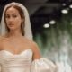 A bride wearing an off-the-shoulder wedding dress with voluminous sleeves and a long veil, standing in a softly lit room. The dress features a structured bodice with elegant folds.