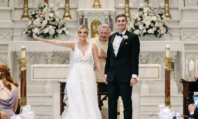 A bride and groom stand at the altar, beaming with joy after being pronounced married. The bride wears a lace wedding dress, and the groom is in a black tuxedo.