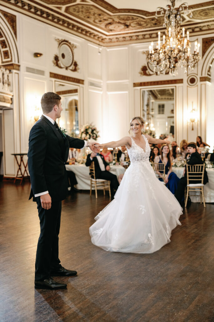 A bride and groom share their first dance in a grand ballroom adorned with chandeliers and elegant décor.