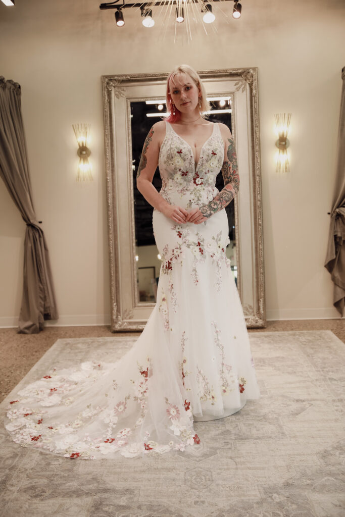 Bride in a detailed floral gown standing in a bridal suite, with elegant décor.